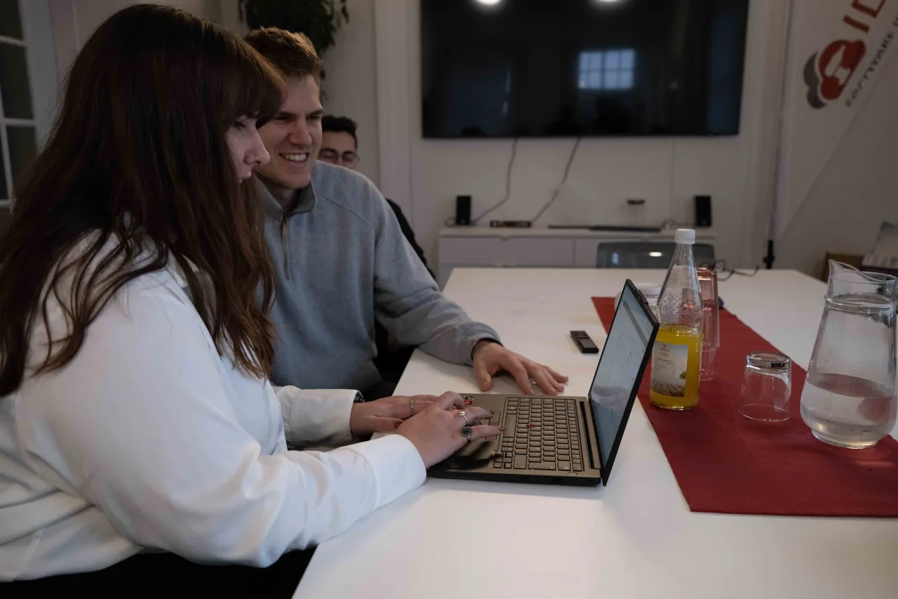 Eine junge Frau und zwei junge Männer sitzen an einem Tisch und zwei arbeiten gemeinsam an einem Laptop. Sie tragen legere Alltagskleidung und haben Getränke vor sich stehen.