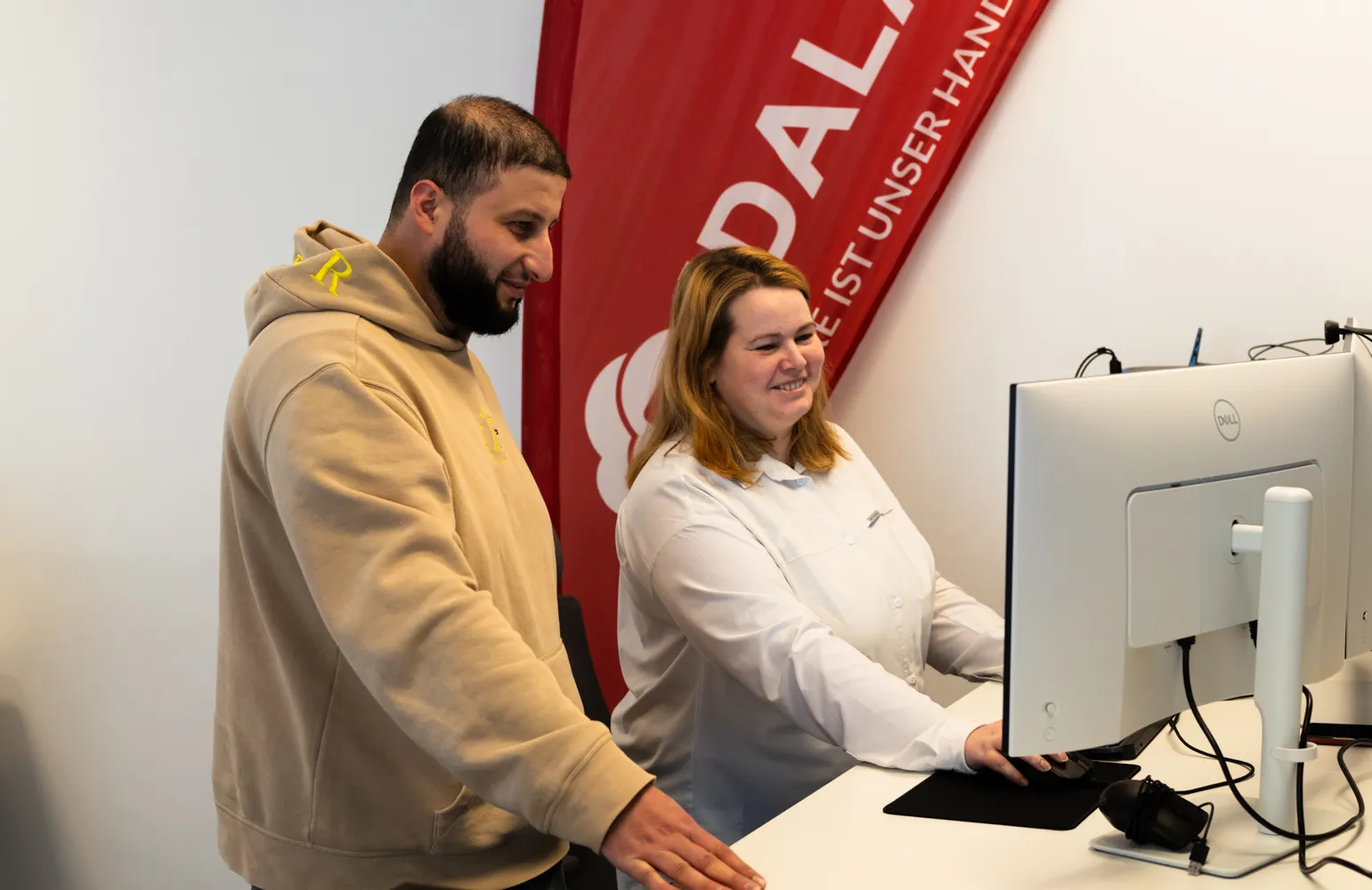Ein junger Mann und eine junge Frau arbeiten gemeinsam an einem Stehschreibtisch und schauen lächelnd auf einen Computerbildschirm. Im Hintergrund ist ein rotes IDALABS-Banner zu sehen