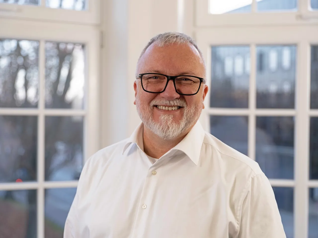 Ein Mann mit braunen Haaren und Dreitagebart lächelt in die Kamera. Er trägt Brille und ein weißes Hemd.