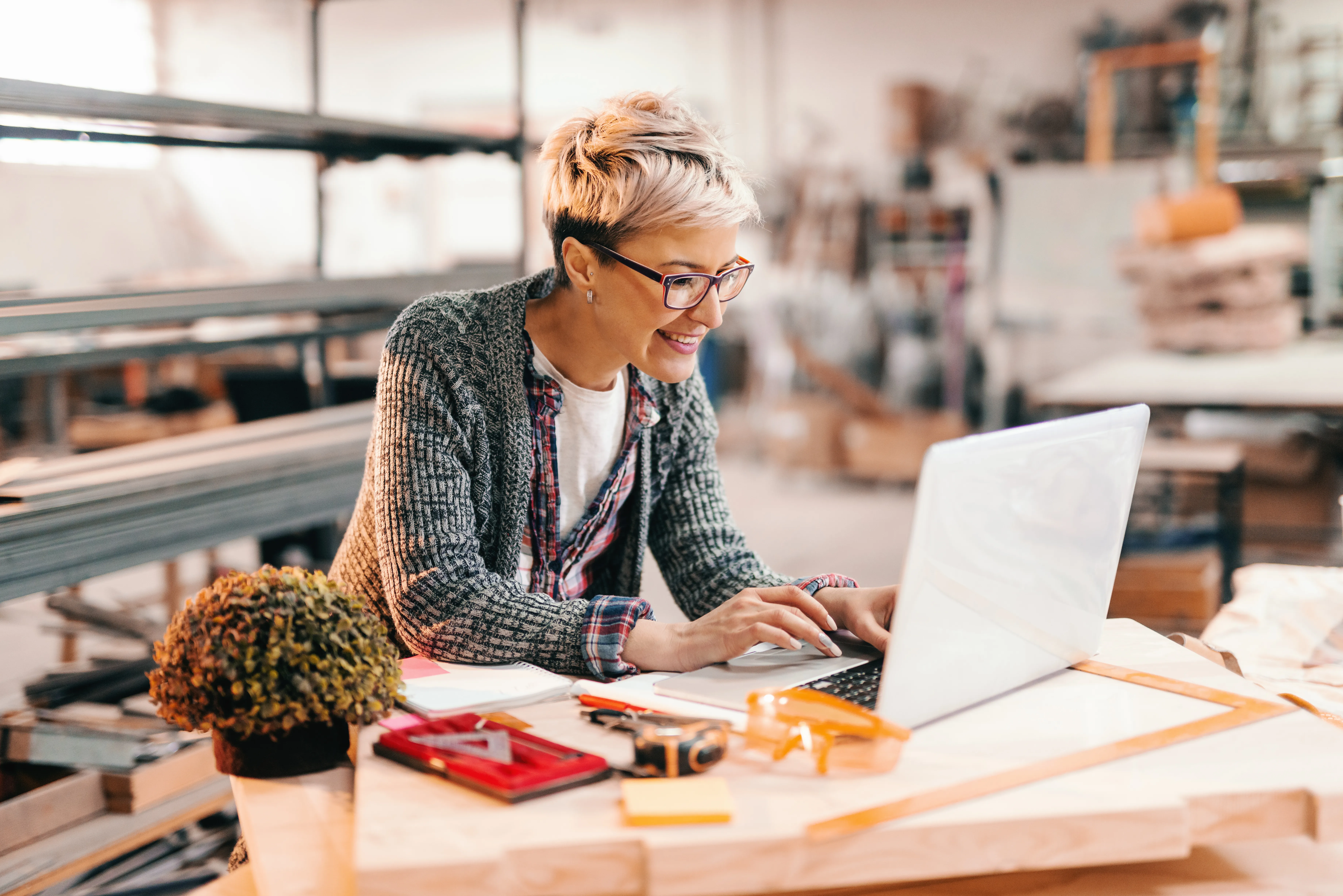 Eine junge Frau mit Kurzhaarschnitt und Brille steht an einem Schreibtisch und arbeitet an einem Laptop. Um sie herum ist verschwommen eine helle Werkstatt zu sehen, in der Holz- und Metallteile sowie verschiedene Werkzeuge angedeutet sind. 