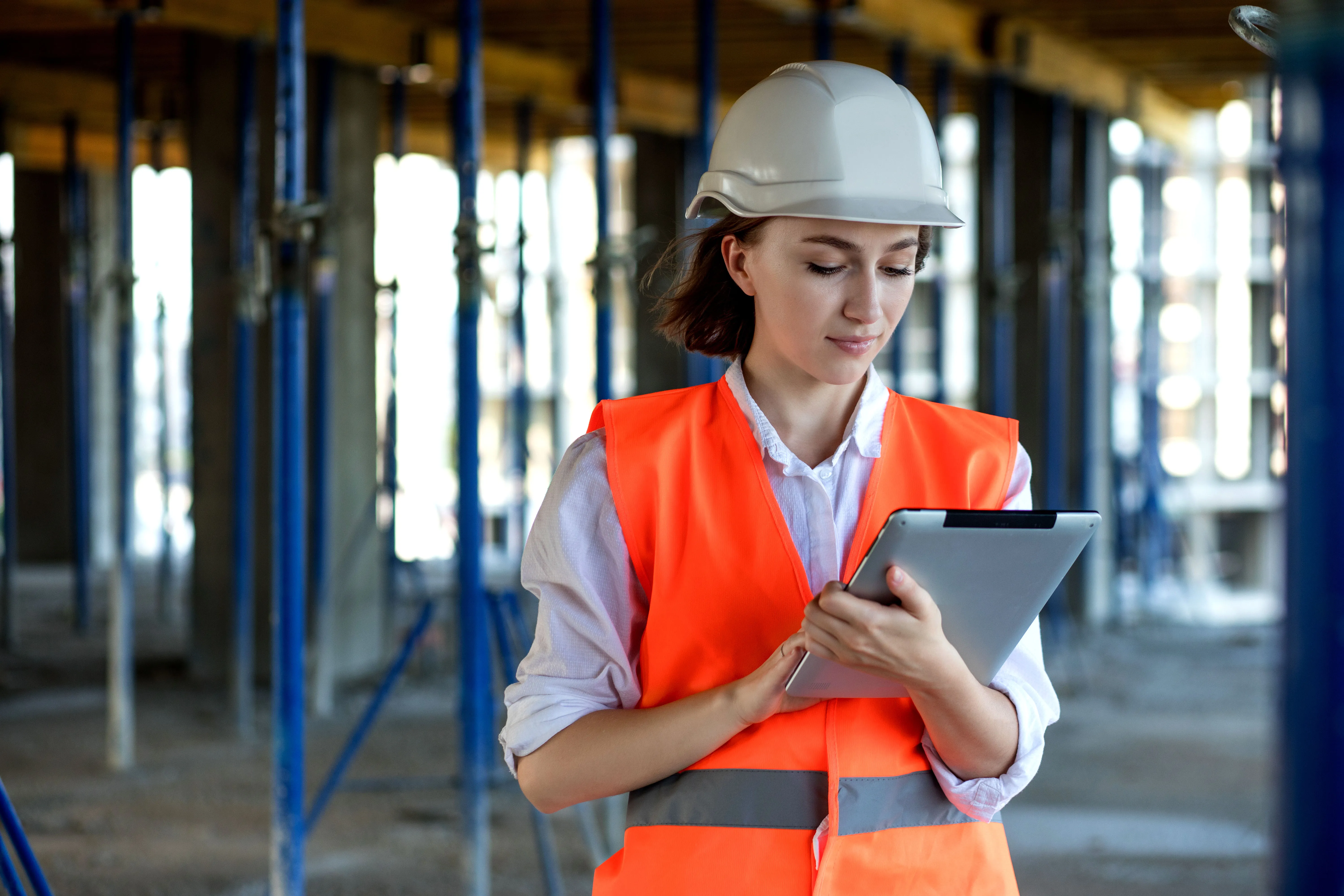  Eine junge Frau, die eine orangefarbene Warnweste und einen weißen Sicherheitshelm trägt, steht vor einer halbfertigen Baukonstruktion. Sie arbeitet an einem Tablet.